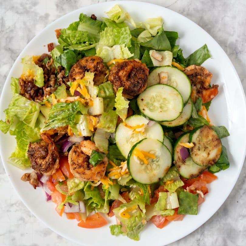 Grilled shrimp salad with lettuce, cucumbers, tomatoes, cheese, and red onions on a white plate.