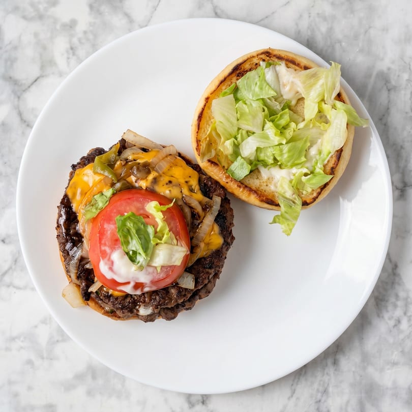 Open-faced burger with melted cheese, grilled onions, tomato, lettuce, and sauce on a white plate.